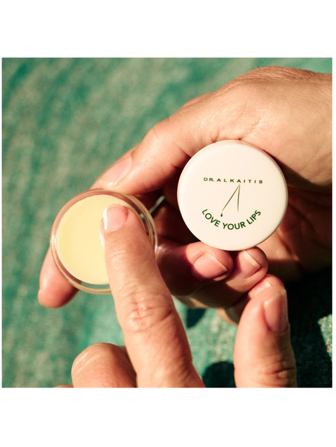 Hand holding Organic Lip Treatment, Love Your Lips, by Dr. Alkaitis against a blurred natural background.