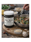 Jar of Raw Black Tahini by Glaser Farms with a spoonful being added to a dish in a kitchen setting.