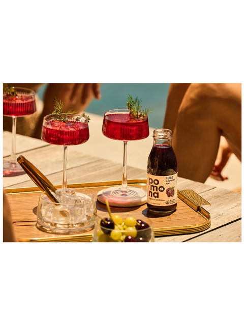 A bottle of Pomona Blackberry Juice on a wooden serving tray next to three pink cocktails in coupe glasses, with a blurred beach setting and people in the background.
