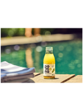 A bottle of Pomna celery juice with a white label and green lemon graphic, filled with yellow-orange liquid, sitting on a wooden deck next to a swimming pool with folded towels in the foreground.