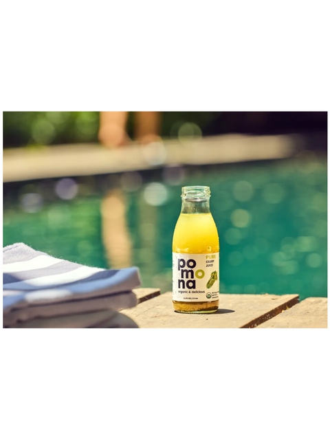 A bottle of Pomna celery juice with a white label and green lemon graphic, filled with yellow-orange liquid, sitting on a wooden deck next to a swimming pool with folded towels in the foreground.