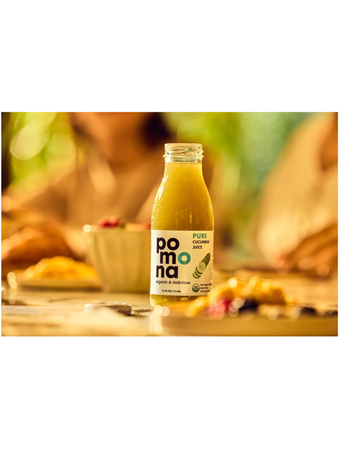 A bottle of Pomona Pure Cucumber Juice standing on a table in a warm, blurred dining setting with people in the background.