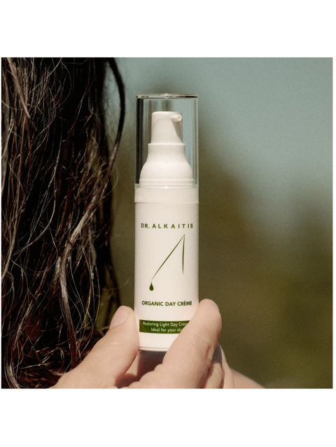 Hand holding a bottle of Dr. Alkaitis Organic Day Creme against a blurred background
