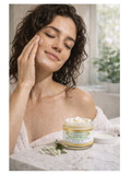 Woman applying From Russia With Love, Regenerating Moisturizer, 2oz, Simply Divine Botanicals to her face with a jar of cream on a table in the foreground.