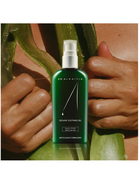 Green bottle of Dr. Alkaitis Organic Soothing Gel held by hands with aloe vera leaves in the background