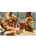 Three people sitting on a wooden deck by a pool with the ocean in the background, sharing red cocktails and a bottle of soda. The scene is brightly lit with warm sunlight.