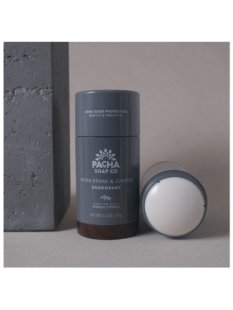 A dark gray cylindrical deodorant stick from Pacha Soap Co stands upright next to its open cap on a neutral surface, with a textured gray block in the background.