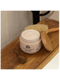Pacha Santal & Amber body scrub in a beige jar, displayed on a wooden tray with a wooden spoon, a natural bristle brush, and its bamboo lid.