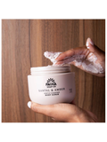 A person's hands holding a jar of Pacha Soap Co Santal & Amber Gentle Cleansing Body Scrub against a warm wood panel background.