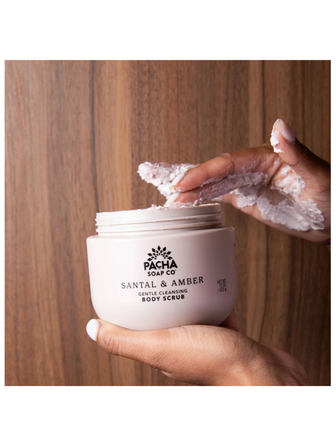 A person's hands holding a jar of Pacha Soap Co Santal & Amber Gentle Cleansing Body Scrub against a warm wood panel background.