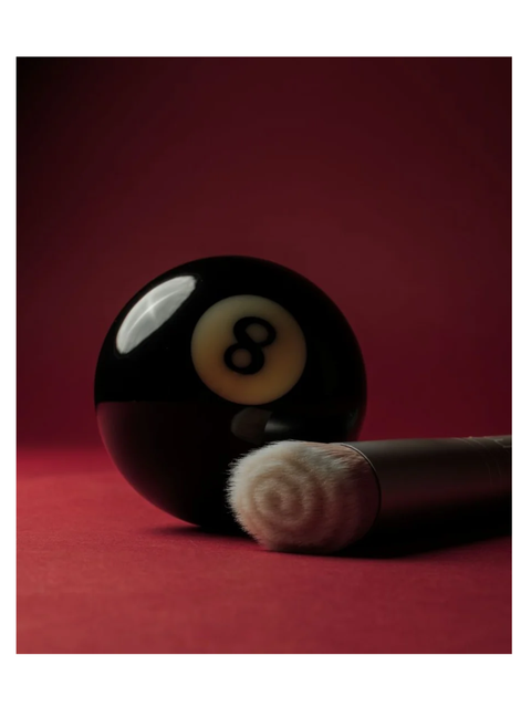 A glossy black 8-ball pool cue resting on a red surface against a dark red background with a skin2skin foundation brush by RMS Beauty.