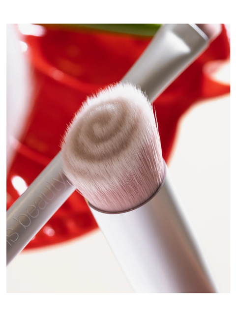 Close-up of skin2skin foundation brush with soft pink bristles and a white handle, with another brush and a blurred red background visible.