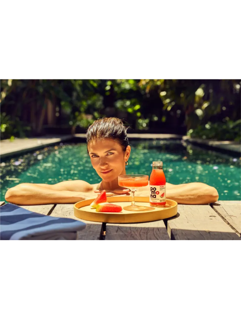 A woman relaxes by a swimming pool with a wooden tray holding a cocktail, watermelon slices, and a bottle of orange-red beverage. The background shows green foliage and the pool water.