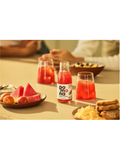 A bottle of Pomona juice with two clear glasses filled with the red beverage, set on a table with snacks and watermelon slices in a warm, softly blurred dining setting.