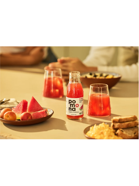A bottle of Pomona juice with two clear glasses filled with the red beverage, set on a table with snacks and watermelon slices in a warm, softly blurred dining setting.