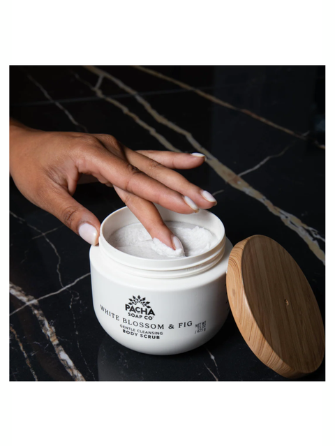 Jar of White Blossom & Fig Body Scrub, Pacha Soap with a hand reaching in to scoop some out, on a marble surface.